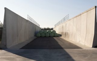 Vista interna di un silo agricolo in cemento armato prefabbricato, con pareti alte dotate di ringhiere di sicurezza. All'interno sono disposte balle di insilato avvolte in plastica verde.