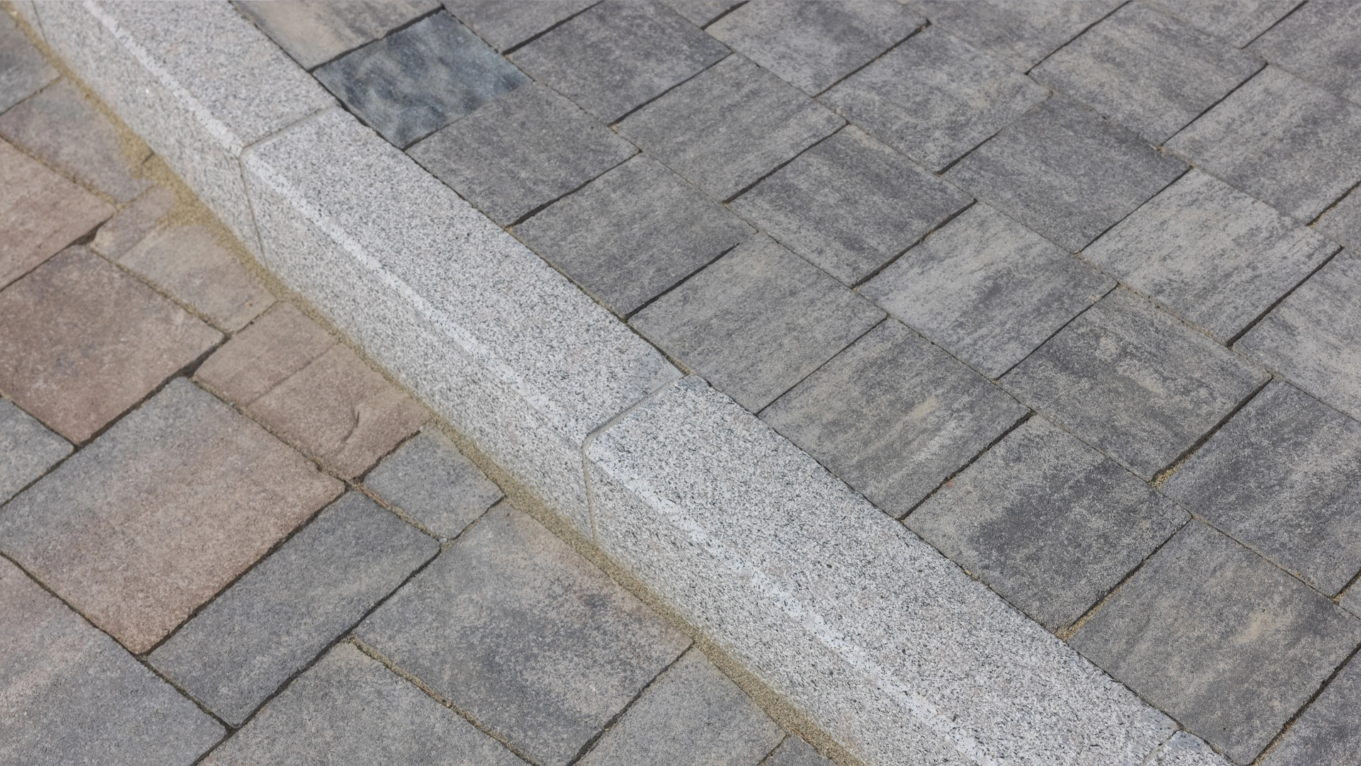 Cordolo stradale in granito con finitura grezza, separa due superfici pavimentate con mattoni autobloccanti di tonalità diverse.