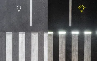 Confronto tra un attraversamento pedonale tradizionale e uno illuminato a LED. A sinistra, le strisce pedonali appaiono normali, mentre a destra sono evidenziate da una luce bianca intensa. Un'icona di lampadina spenta è presente nella parte sinistra, mentre nella destra è accesa e gialla.