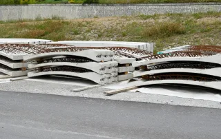 Pannelli prefabbricati in calcestruzzo con armatura metallica accatastati in un'area di cantiere. Gli elementi curvi, destinati al rivestimento di una galleria, sono disposti in pile e separati.
