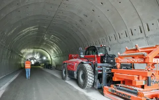 Interno di un tunnel in costruzione, con pareti rivestite da segmenti prefabbricati in calcestruzzo. Un operaio con gilet arancione cammina lungo la galleria, mentre un grande veicolo rosso con attrezzature marcate 'GREEN POWER' è parcheggiato sul lato destro.