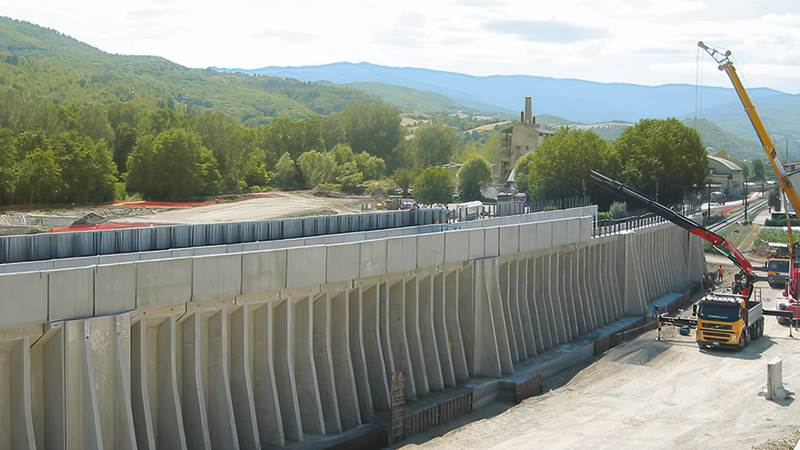 Costruzione di una galleria scatolare in cemento armato con pareti rinforzate da elementi inclinati di sostegno. Sul cantiere sono presenti camion, gru e operai impegnati nei lavori di montaggio.