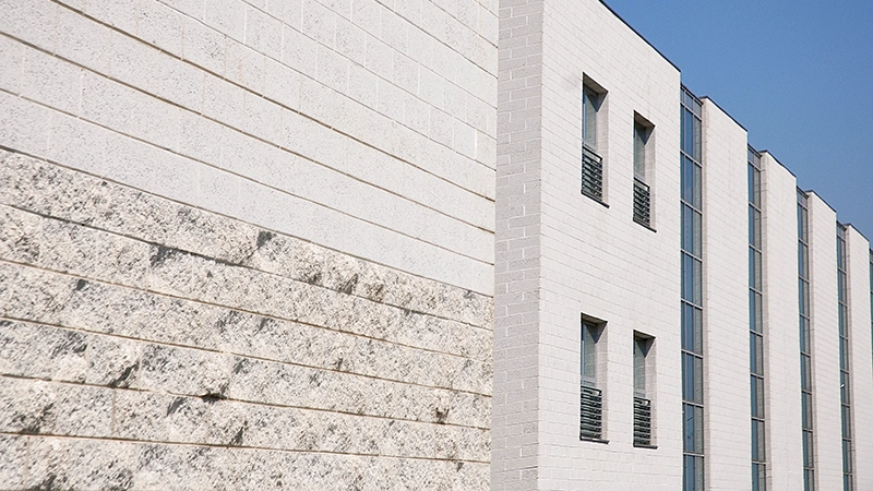 Edificio con pareti in blocchi di cemento di diverse finiture, alcune lisce e altre ruvide e testurizzate. La struttura presenta una combinazione di superfici chiare e sezioni vetrate verticali con infissi metallici.