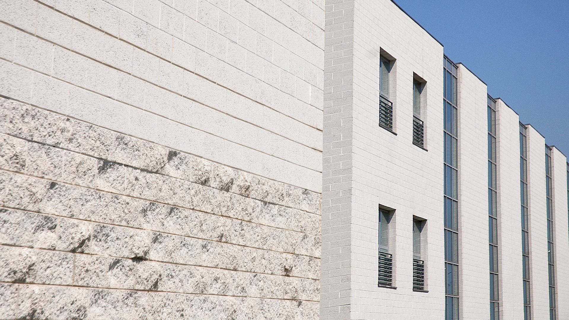 Edificio con pareti in blocchi di cemento di diverse finiture, alcune lisce e altre ruvide e testurizzate. La struttura presenta una combinazione di superfici chiare e sezioni vetrate verticali con infissi metallici.