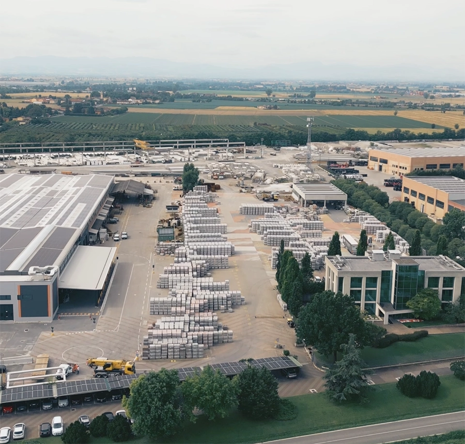"Vista aerea dello stabilimento dell'azienda Paver, con capannoni industriali, uffici e magazzini. Nell'area esterna si notano grandi quantità di materiali impilati, mezzi di trasporto e macchinari. Lo stabilimento è circondato da aree verdi e campi coltivati, con un paesaggio collinare sullo sfondo.