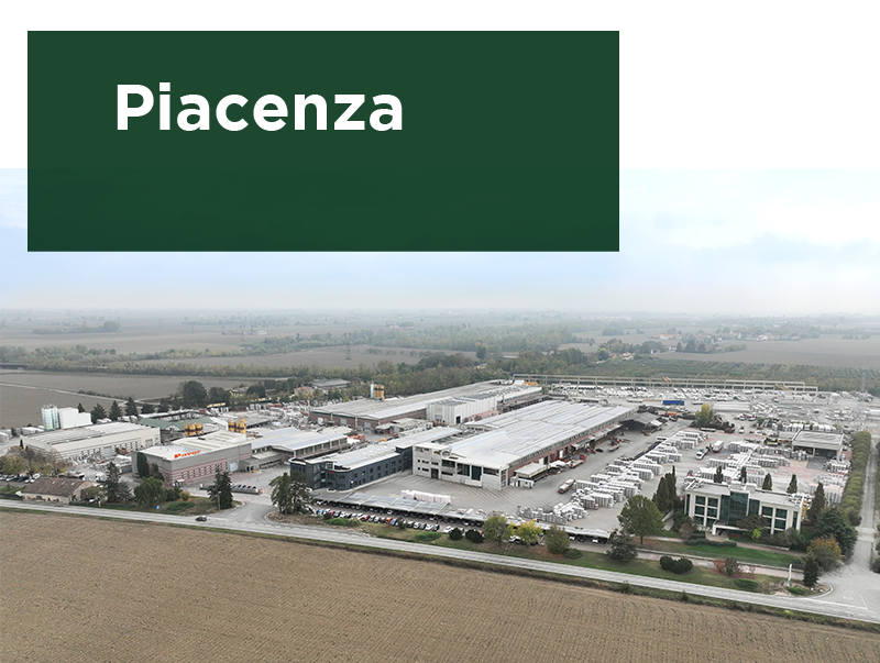 Vista aerea dello stabilimento Paver a Piacenza, con edifici industriali, aree di stoccaggio materiali e parcheggi, circondato da campi agricoli. Testo 'Piacenza' su sfondo verde.