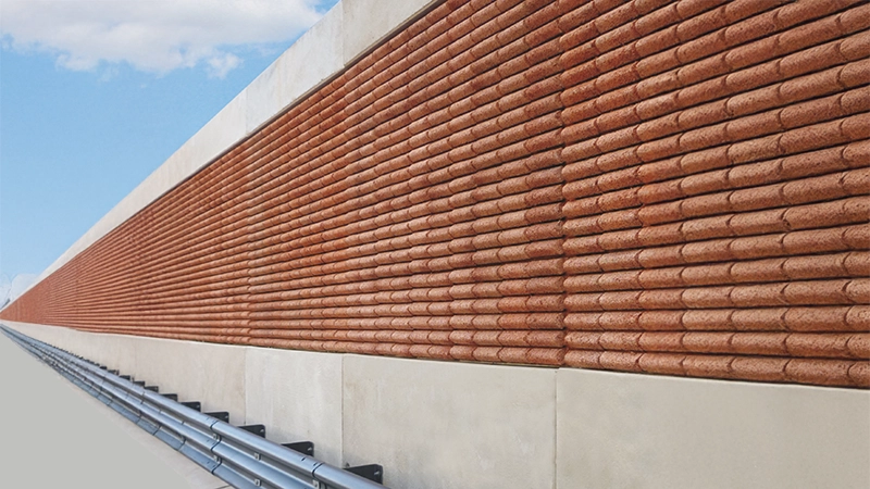 Primo piano di una barriera fonoassorbente lungo una strada, caratterizzata da una texture in rilievo con elementi in laterizio e una base in cemento.