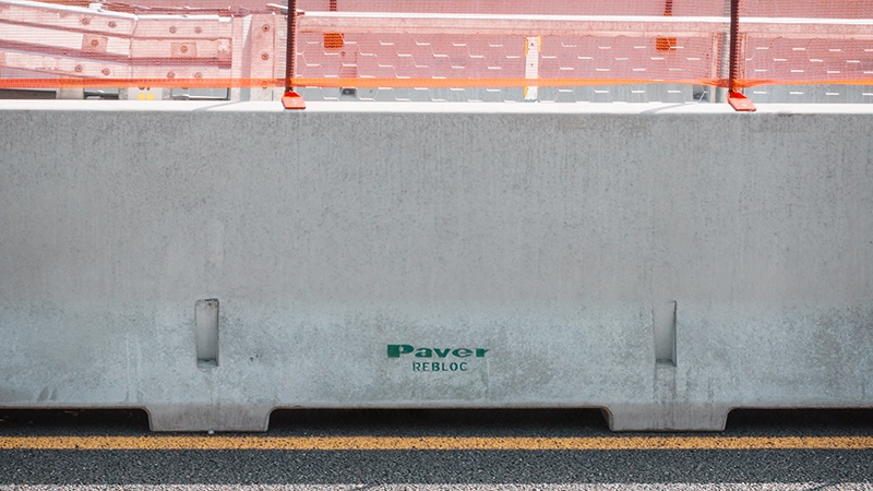 Barriera stradale in calcestruzzo modello New Jersey con marchio 'Paver REBLOC', posizionata lungo una carreggiata con asfalto e segnaletica orizzontale, delimitata da una rete arancione di sicurezza.