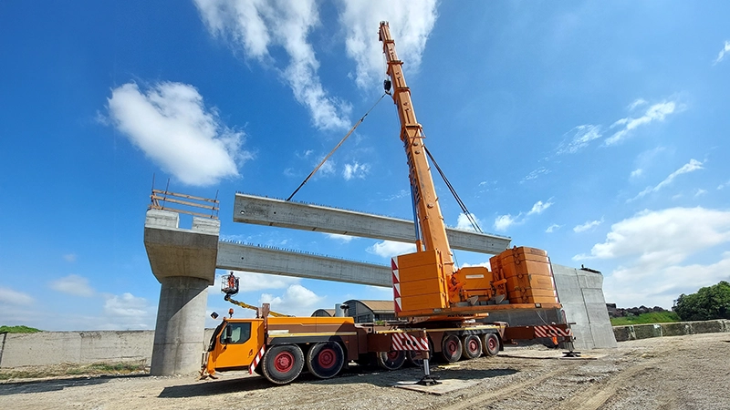 Gru mobile di colore arancione in azione durante l'installazione di travi in cemento armato per la costruzione di un ponte stradale.
