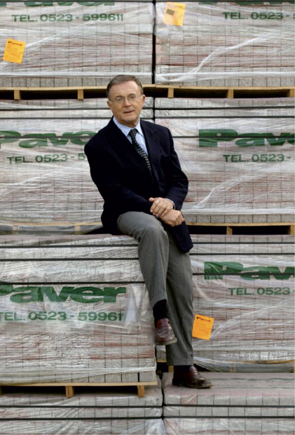 Il fondatore dell'azienda, un uomo anziano vestito elegantemente con giacca scura, camicia e cravatta, è seduto su una pila di materiali da costruzione imballati, con il logo "Paver" ben visibile. Sullo sfondo si notano altre confezioni simili, impilate ordinatamente.