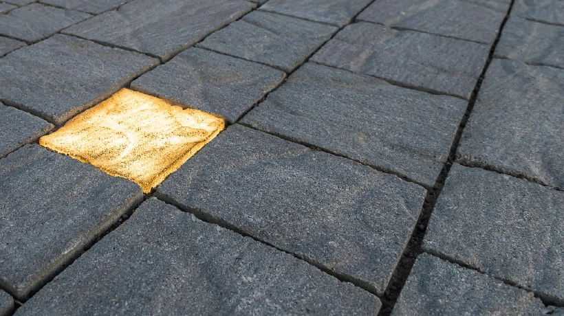 L'immagine mostra una superficie pavimentata con blocchi di pietra di colore grigio scuro, disposti in modo regolare. Tra questi, un singolo blocco si distingue perché è di colore dorato e sembra illuminato, creando un contrasto evidente con il resto della pavimentazione.